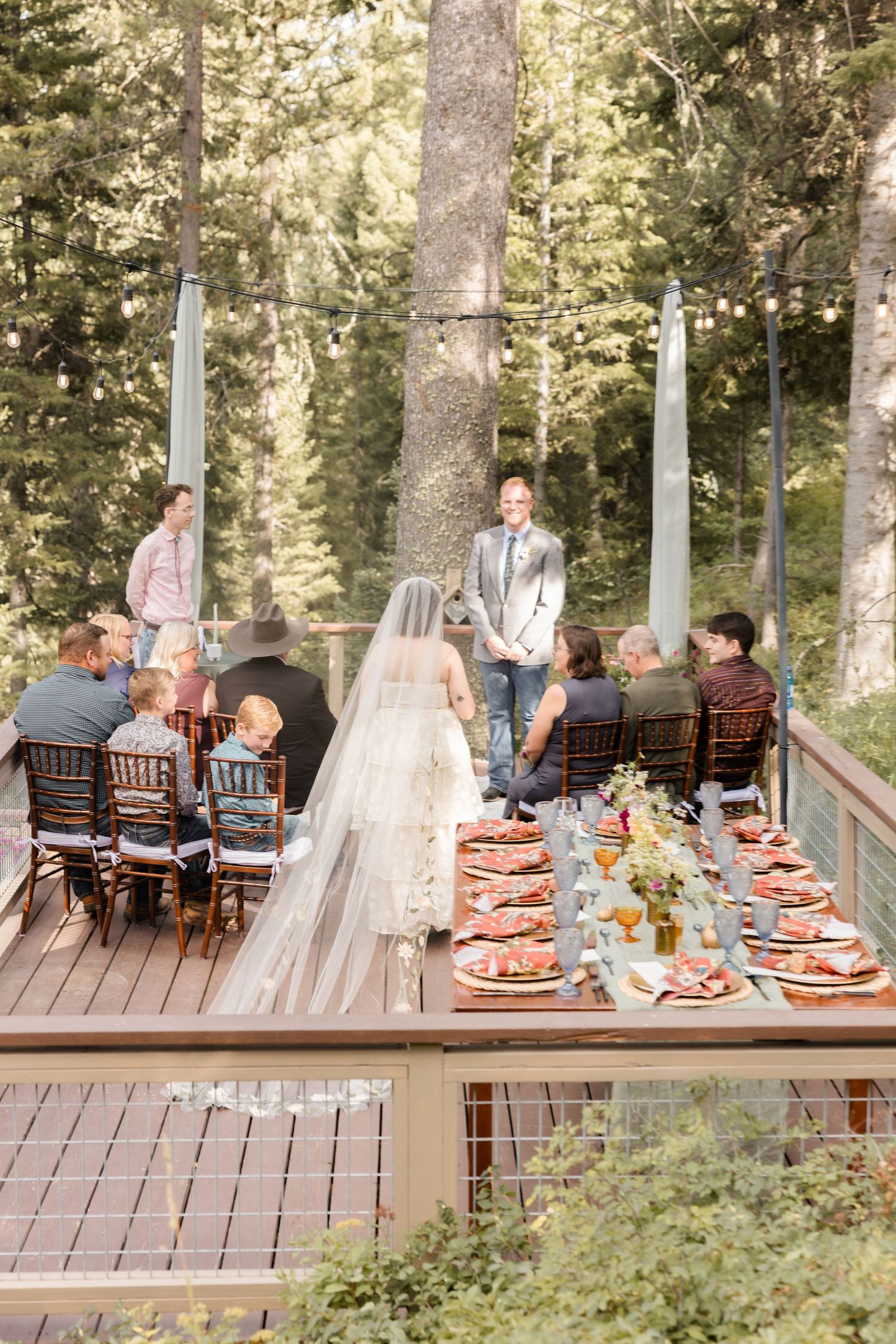 Outdoor forest wedding ceremony on a wooden deck with long dining table, bride in veil, groom, and seated guests among tall trees.
