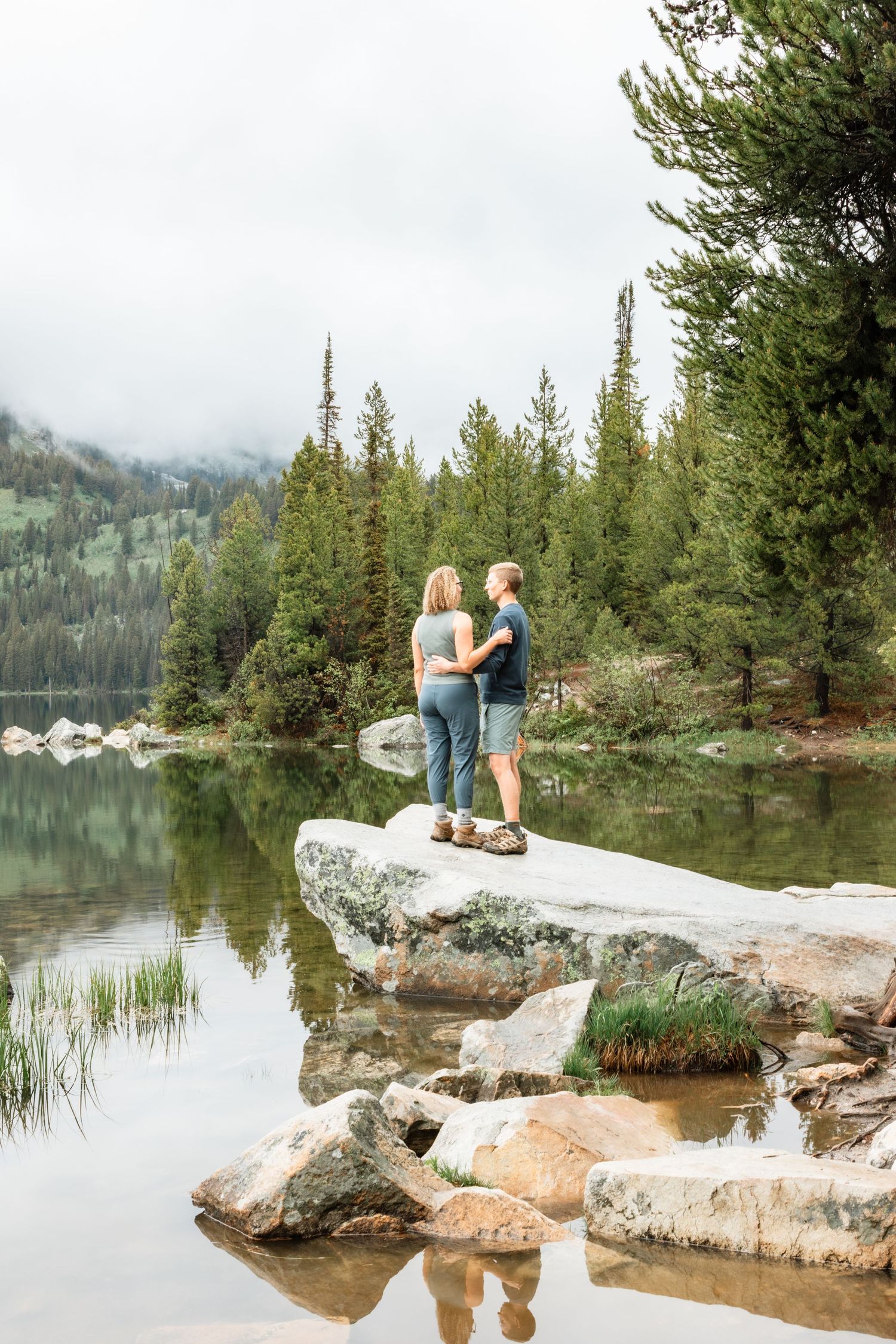 Couple standing on rocks by a serene mountain lake surrounded by evergreen trees and misty peaks.