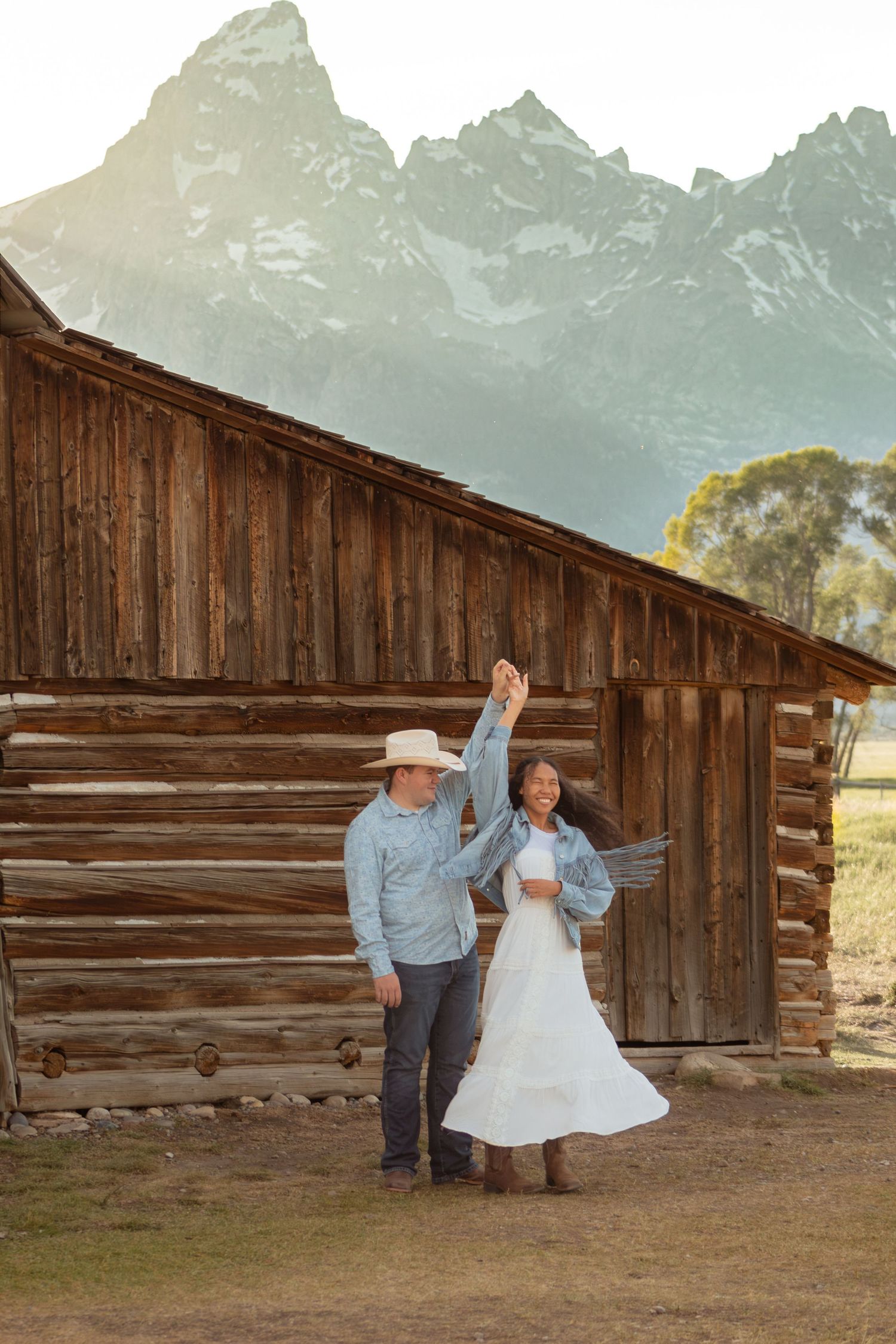 Couple dancing near rustic log barn with Grand Teton mountains backdrop, man in cowboy hat twirling woman in white dress.
