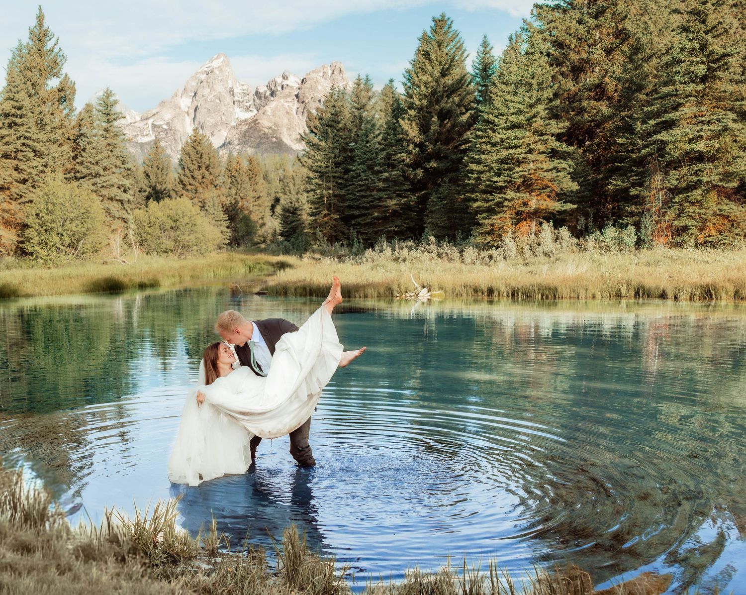 Groom carries bride in white dress while standing in a mountain lake surrounded by pine trees and rocky peaks.