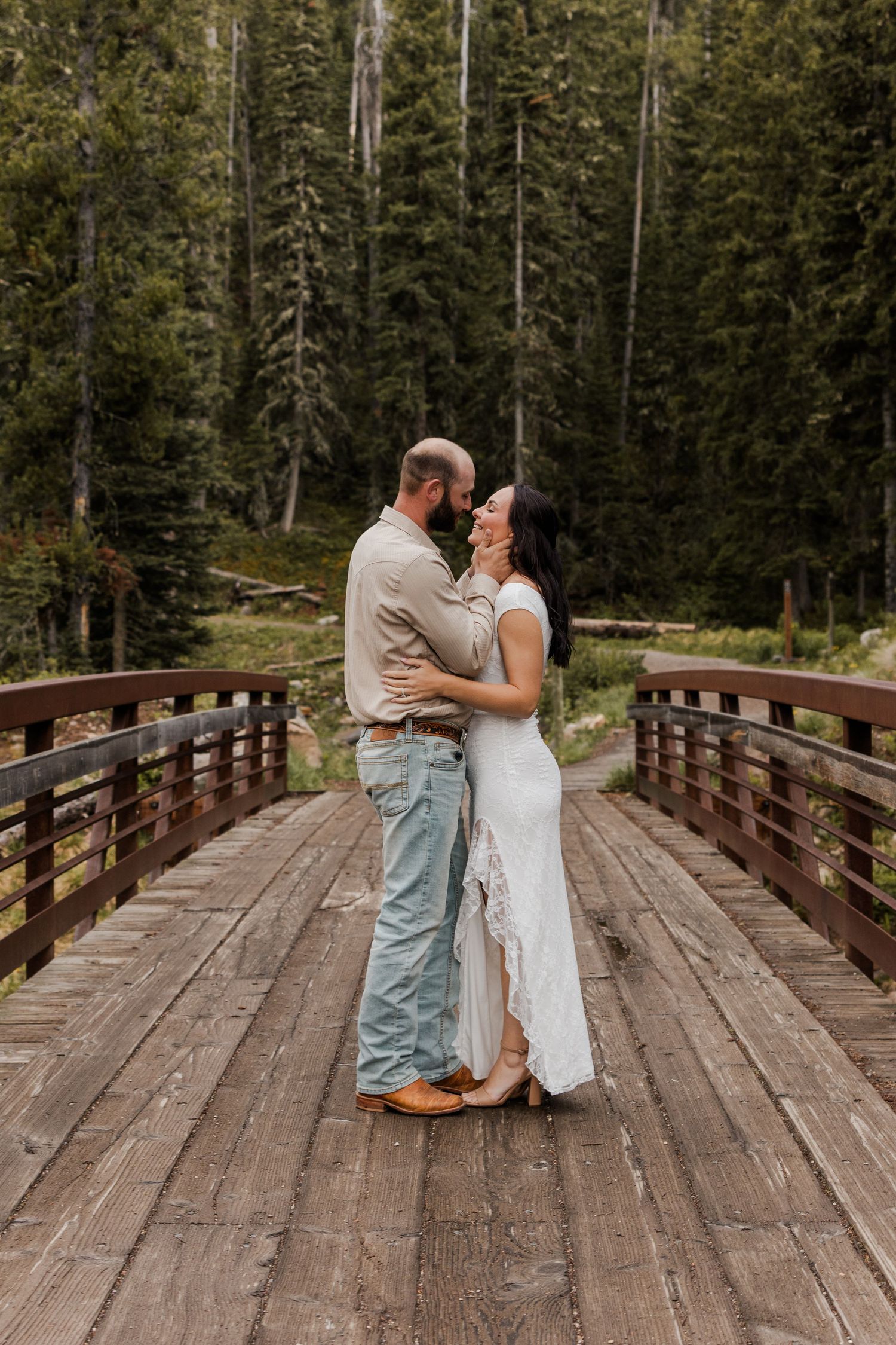 Couple embracing on a wooden bridge surrounded by tall evergreen trees in a lush forest setting.