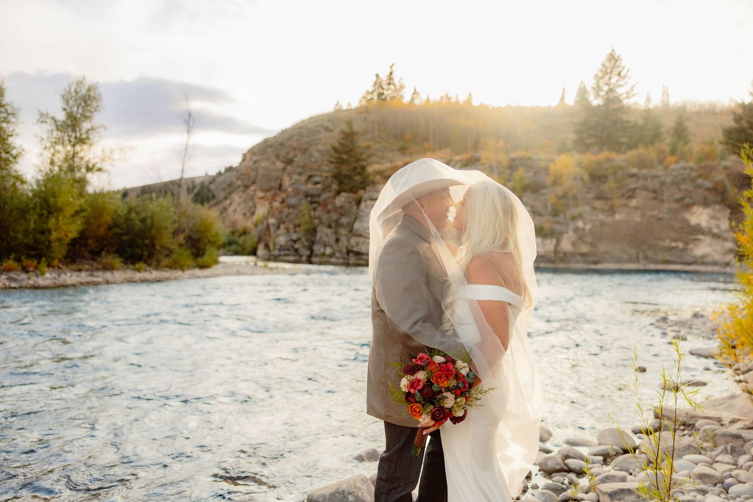 Bride and groom kissing under veil by a mountain river at sunset, bride holding a red bouquet.