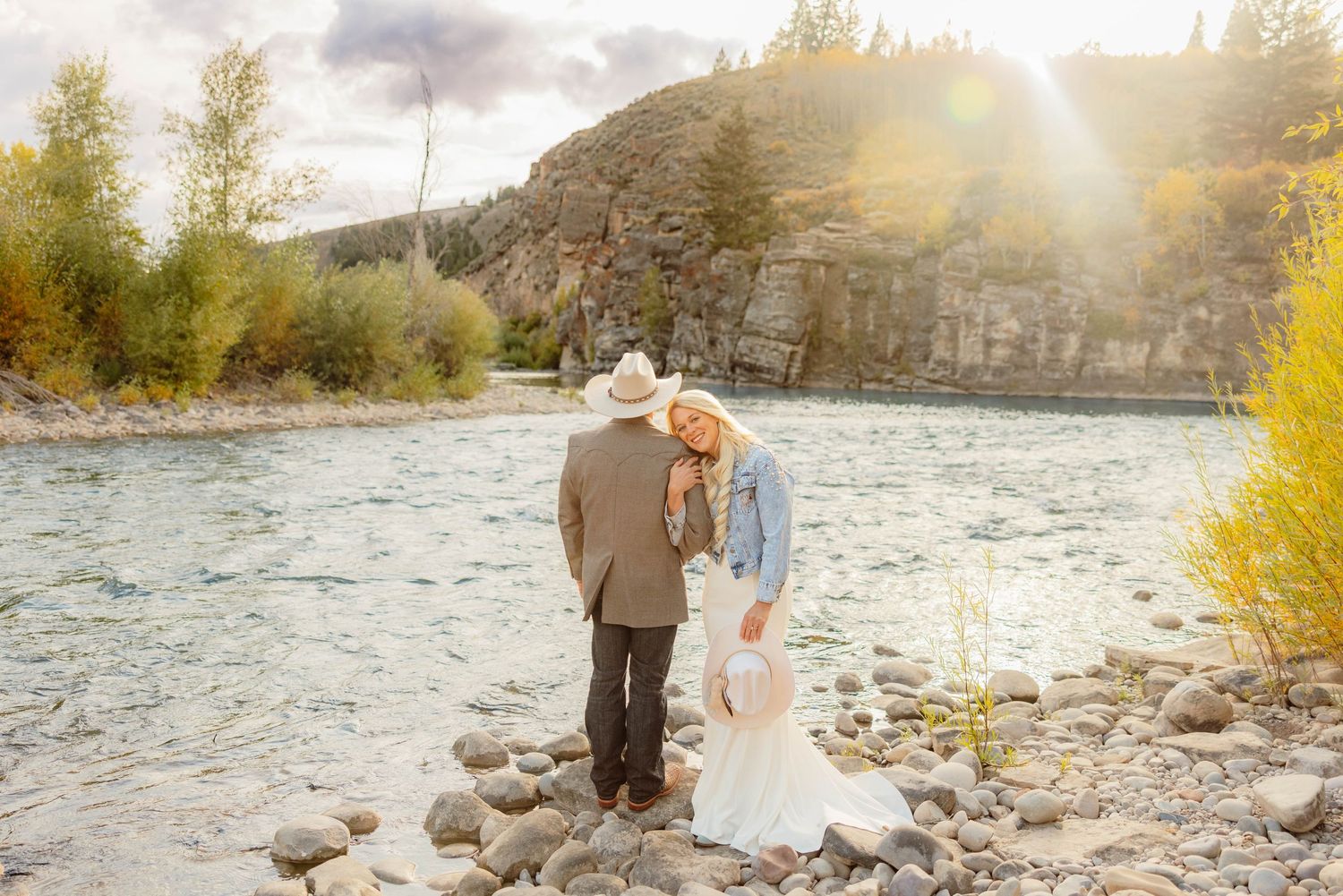 Bride and groom in cowboy hats stand on rocky riverbank with golden sunlight and autumn trees in background.