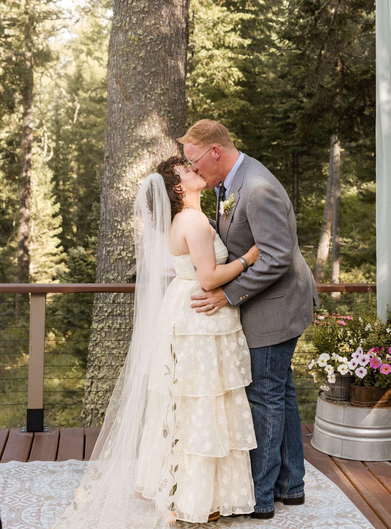 Bride and groom share first kiss in wedding ceremony outdoors on wooden deck surrounded by tall pine trees.