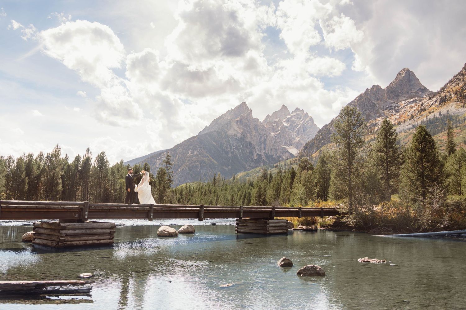 Bride in white dress stands on wooden bridge over mountain river with Grand Teton peaks and pine trees in background.