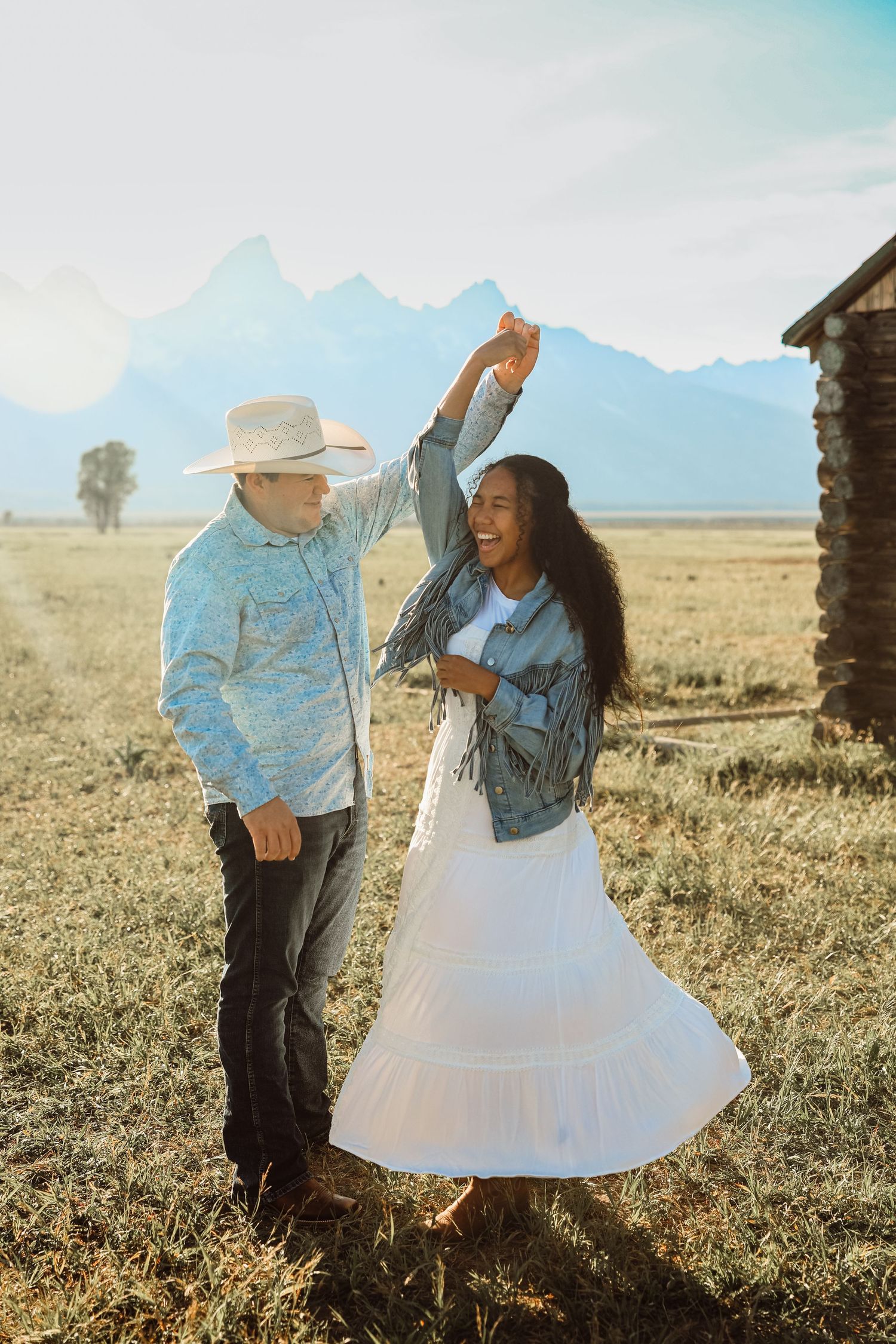 Smiling couple dancing in a grassy field near a rustic log cabin with snow-capped mountains behind, the woman twirling in a white skirt.