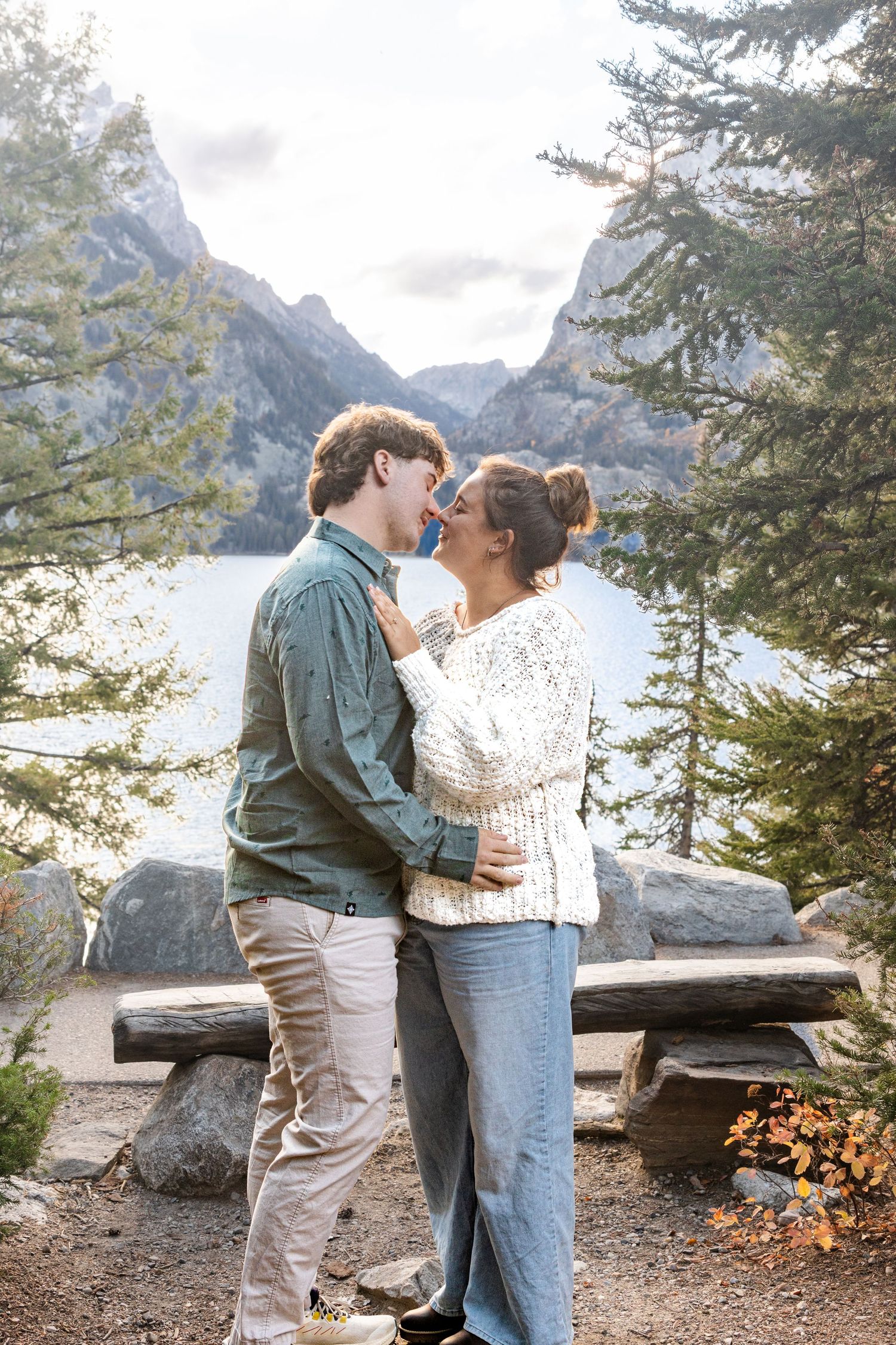 Couple sharing a romantic kiss outdoors near a mountain lake, surrounded by pine trees and rocky peaks.