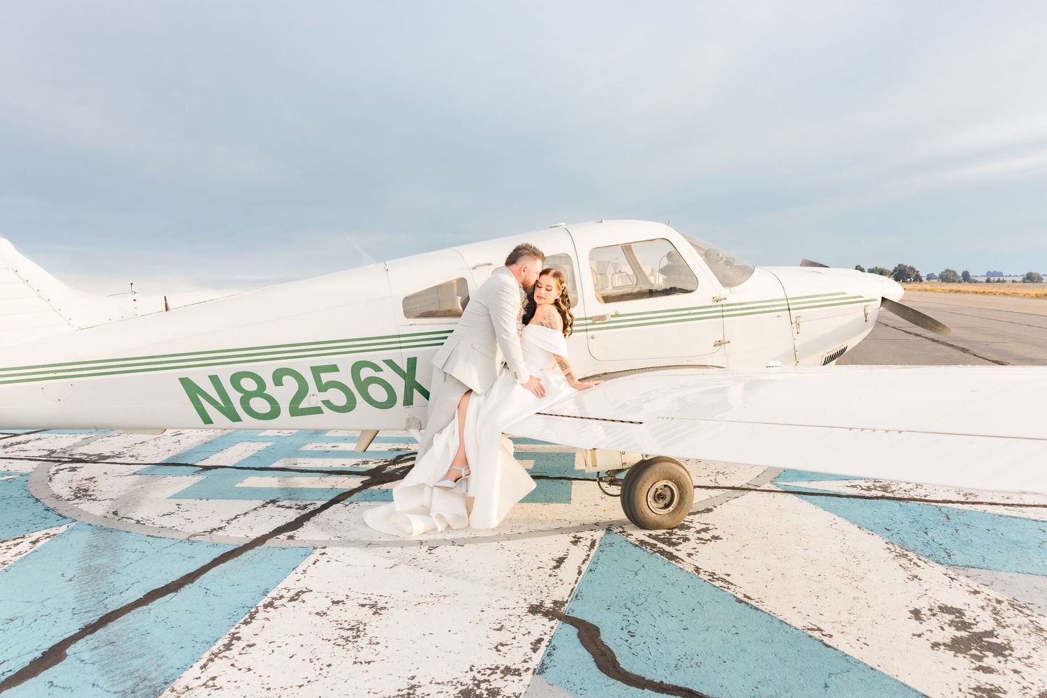 A bride and groom pose romantically in front of a white Piper aircraft on a sunlit airport tarmac.