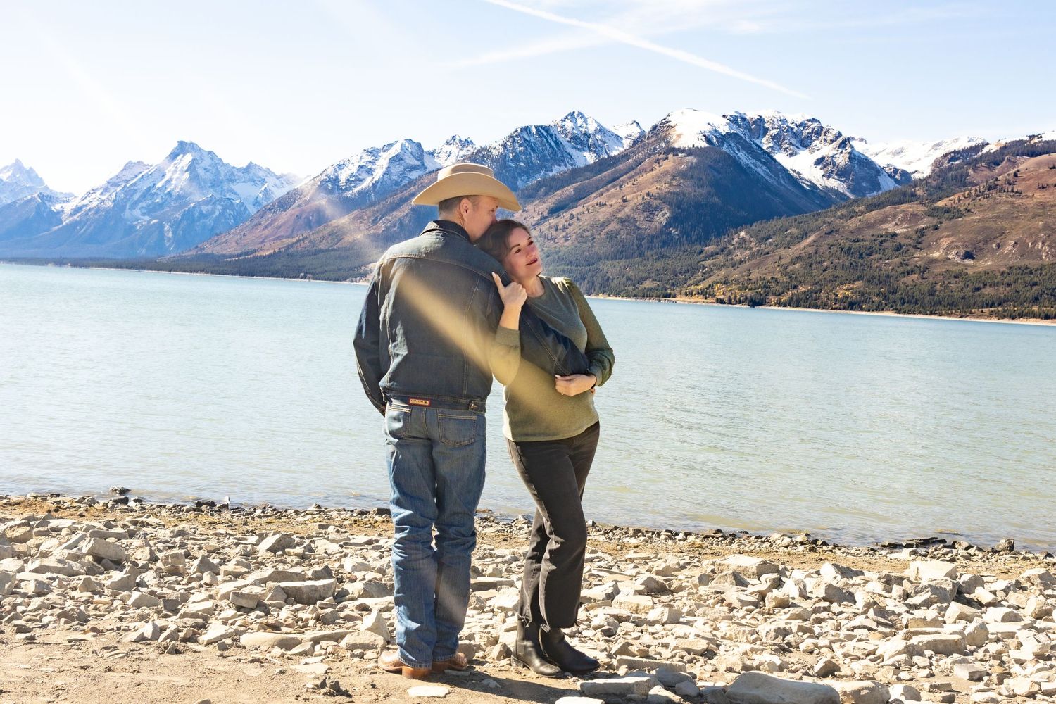 Couple standing by a rocky lakeside with snow-capped mountains in the background on a sunny day.