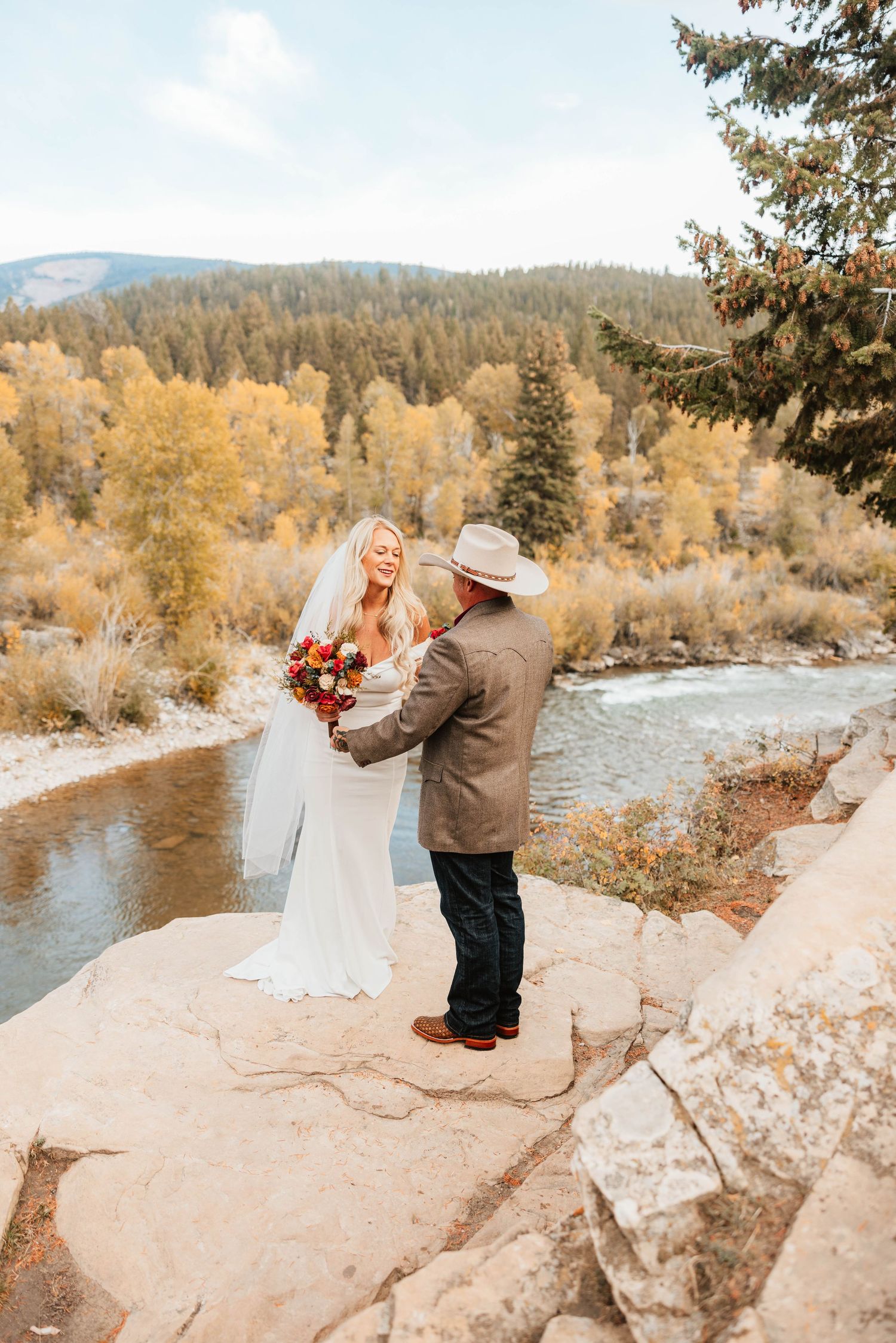 Bride and groom stand on rocky riverbank surrounded by golden autumn trees during outdoor fall wedding ceremony.