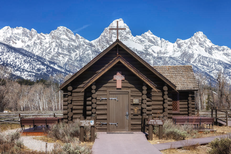 Log cabin chapel with wooden cross atop roof, set against snow-capped Grand Teton mountains under clear blue sky.