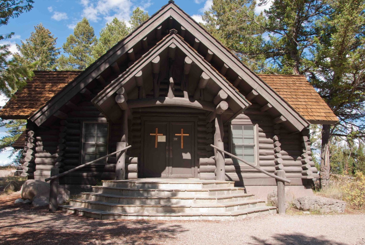 Rustic log cabin chapel with wooden cross on door, surrounded by pine trees under a blue sky.