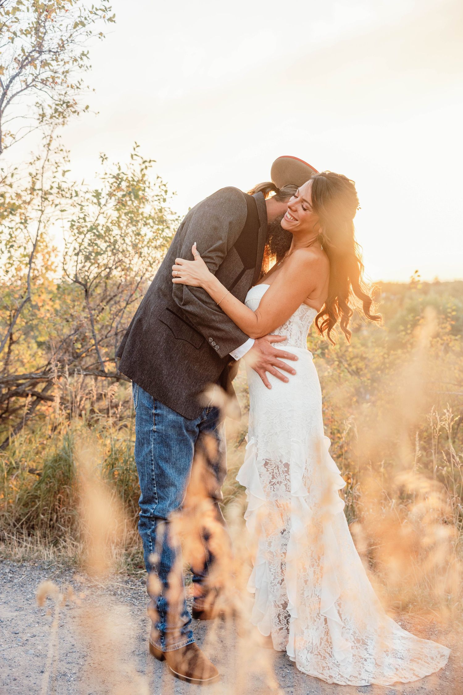 A couple embraces at golden hour, bride in lace gown and groom in cowboy hat, surrounded by wild desert brush.