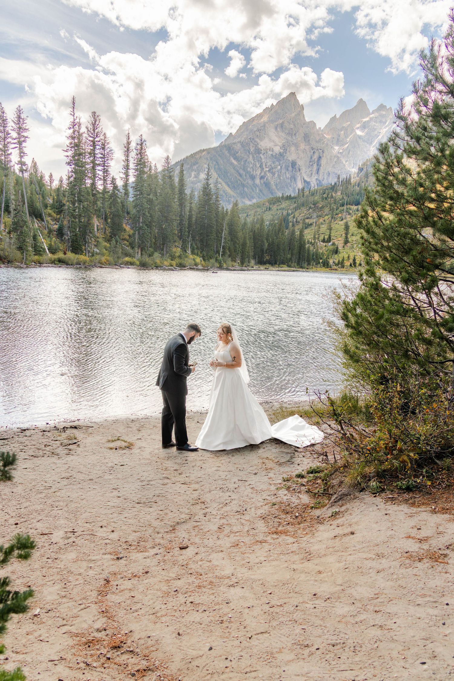 Bride and groom exchange vows on sandy shore of mountain lake with Grand Teton peaks and pine forest backdrop.