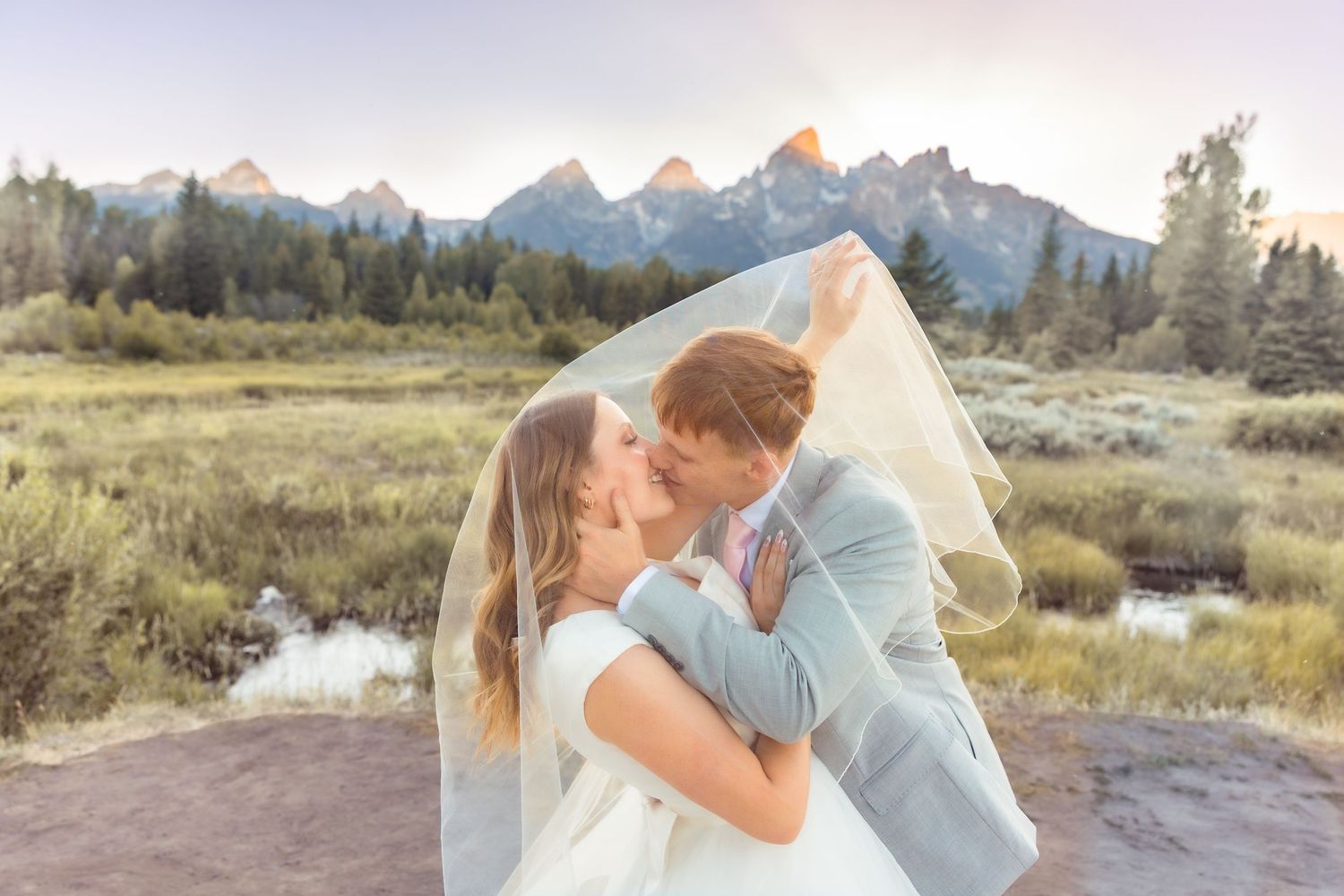 Bride and groom kissing under a flowing veil with Grand Teton mountains in the background at sunset.