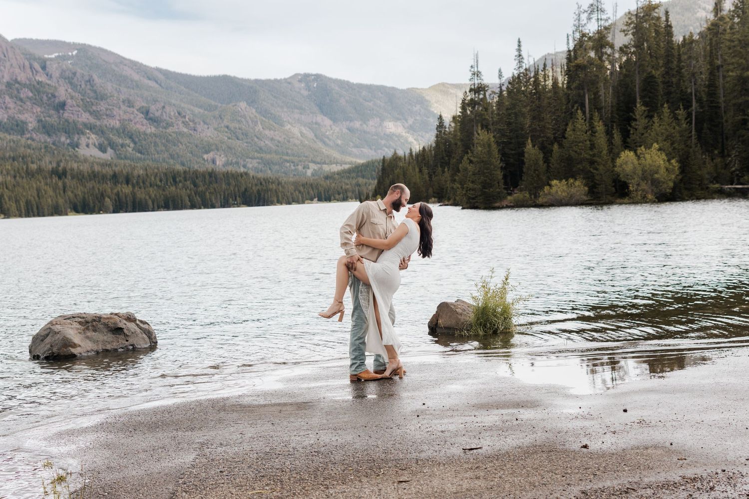 Couple sharing a romantic embrace at a mountain lake shore, surrounded by pine trees and scenic peaks.