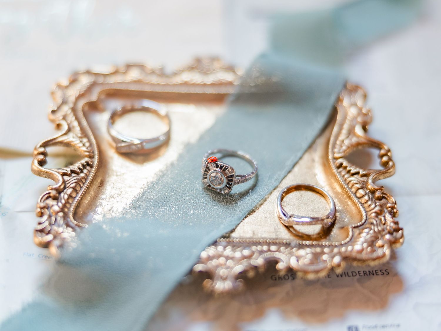 Vintage engagement ring and two wedding bands on an ornate gold tray draped with a dusty blue satin ribbon.