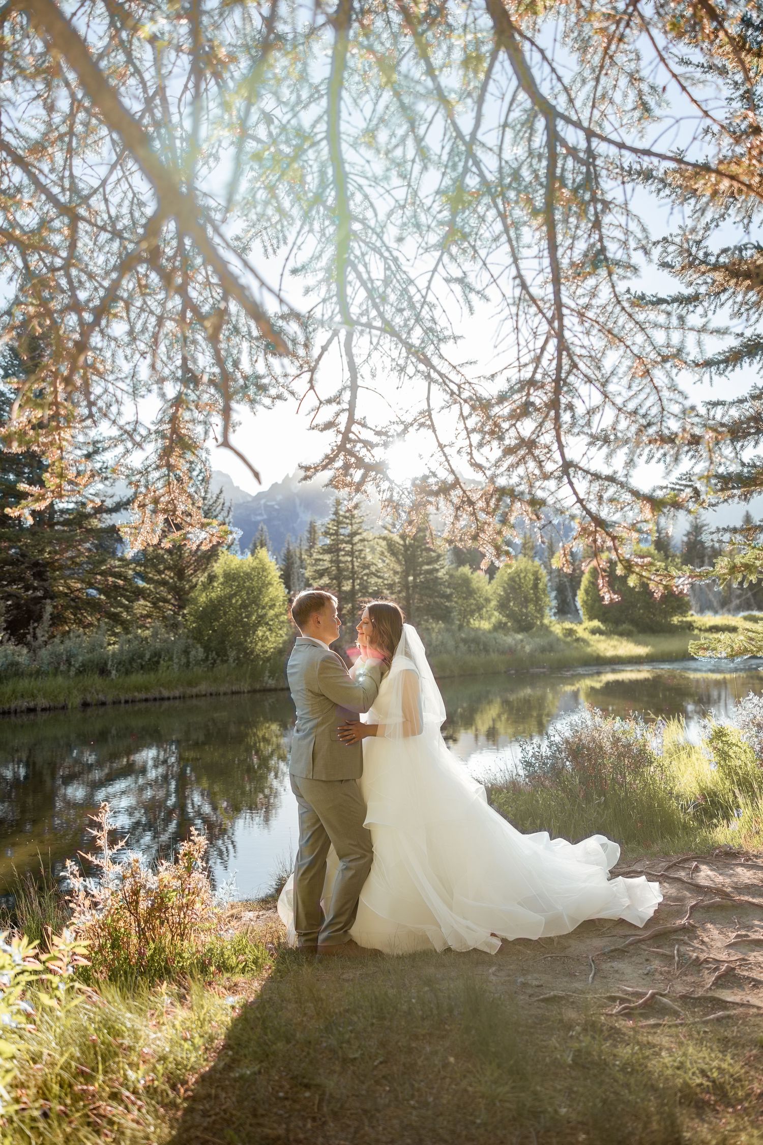 Bride and groom embracing near a mountain lake under sun-dappled pine trees in a romantic outdoor wedding setting.