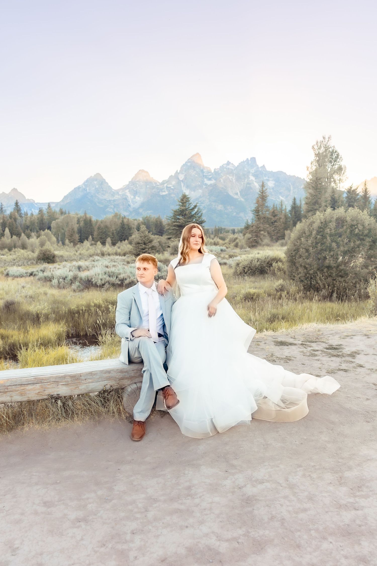 Bride in a flowing white gown and groom in a light gray suit sit on a wooden bench with jagged mountains and golden meadow behind them.