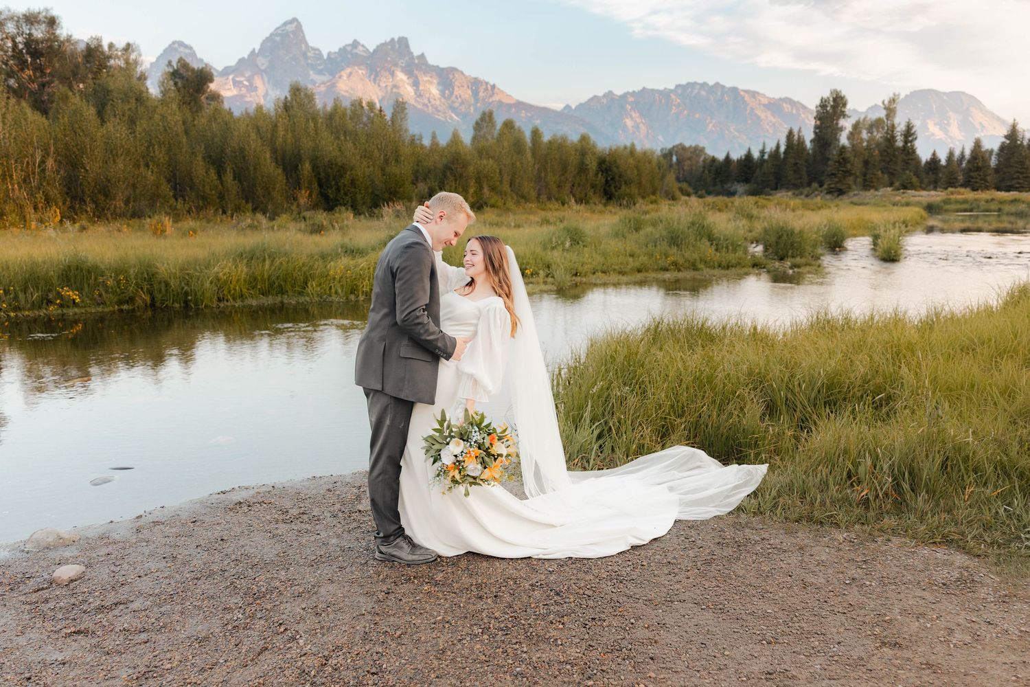 Bride and groom share a romantic kiss by a mountain river in Grand Teton National Park, bride holding a wildflower bouquet.