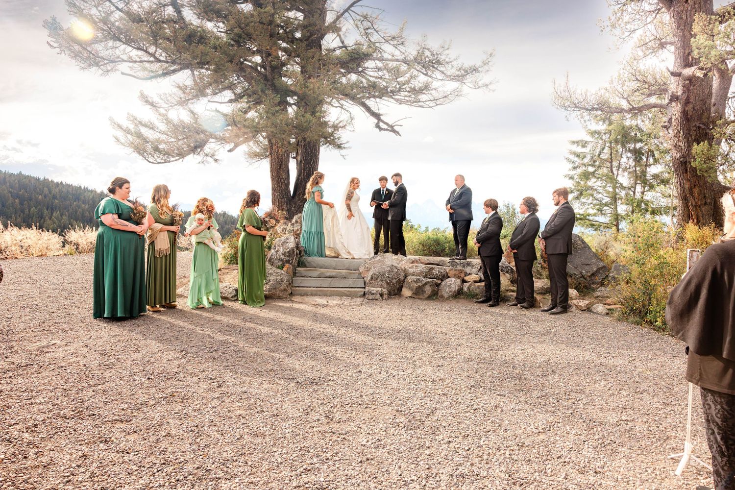 Outdoor wedding ceremony with bridesmaids in green dresses and groomsmen on a gravel terrace surrounded by pine trees.