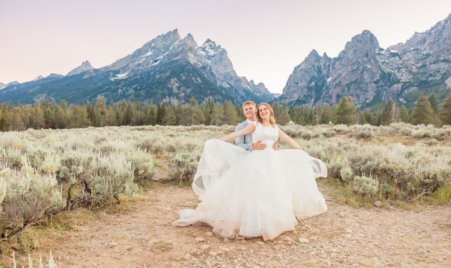 Groom lifts bride in white gown at Grand Teton National Park with mountain peaks and sagebrush backdrop.