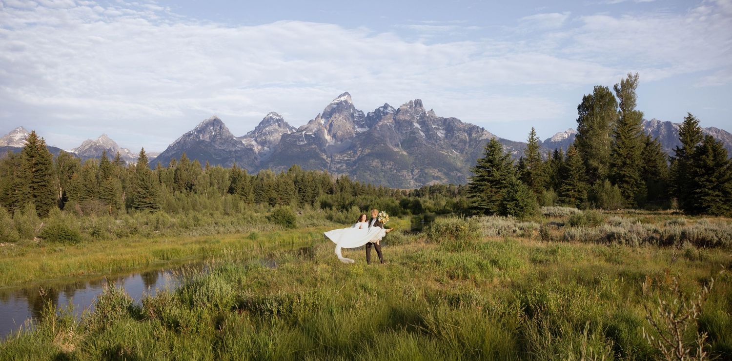 Bride in white dress twirling in lush green meadow with Grand Teton mountain range backdrop under blue sky.