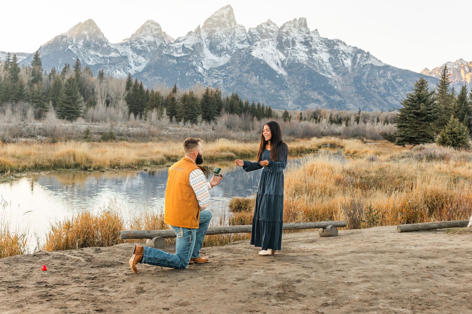 Man proposes on one knee to woman near mountain lake at Grand Teton National Park during golden hour.
