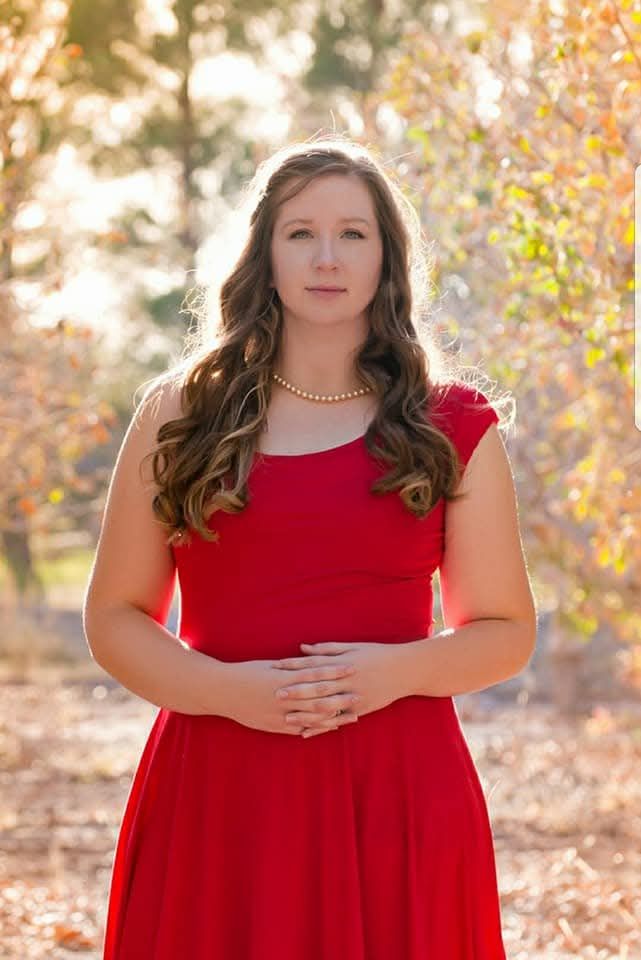 Woman with long wavy brown hair wearing a red dress and pearl necklace stands with hands clasped in a sunlit autumn park.