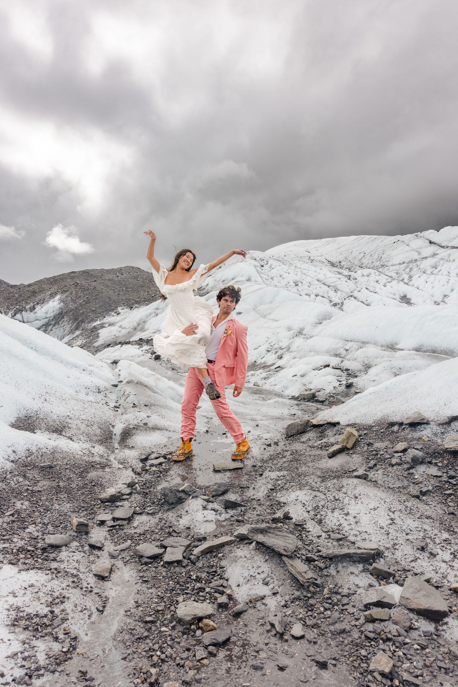 Couple in wedding attire dancing on a glacier, bride in white dress and groom in pink suit with yellow boots.