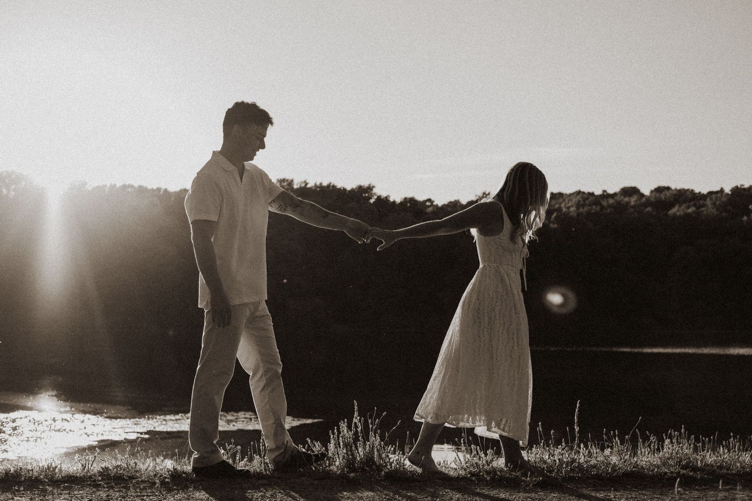 Black and white romantic photo of couple holding hands by a sunlit lake in an open field.