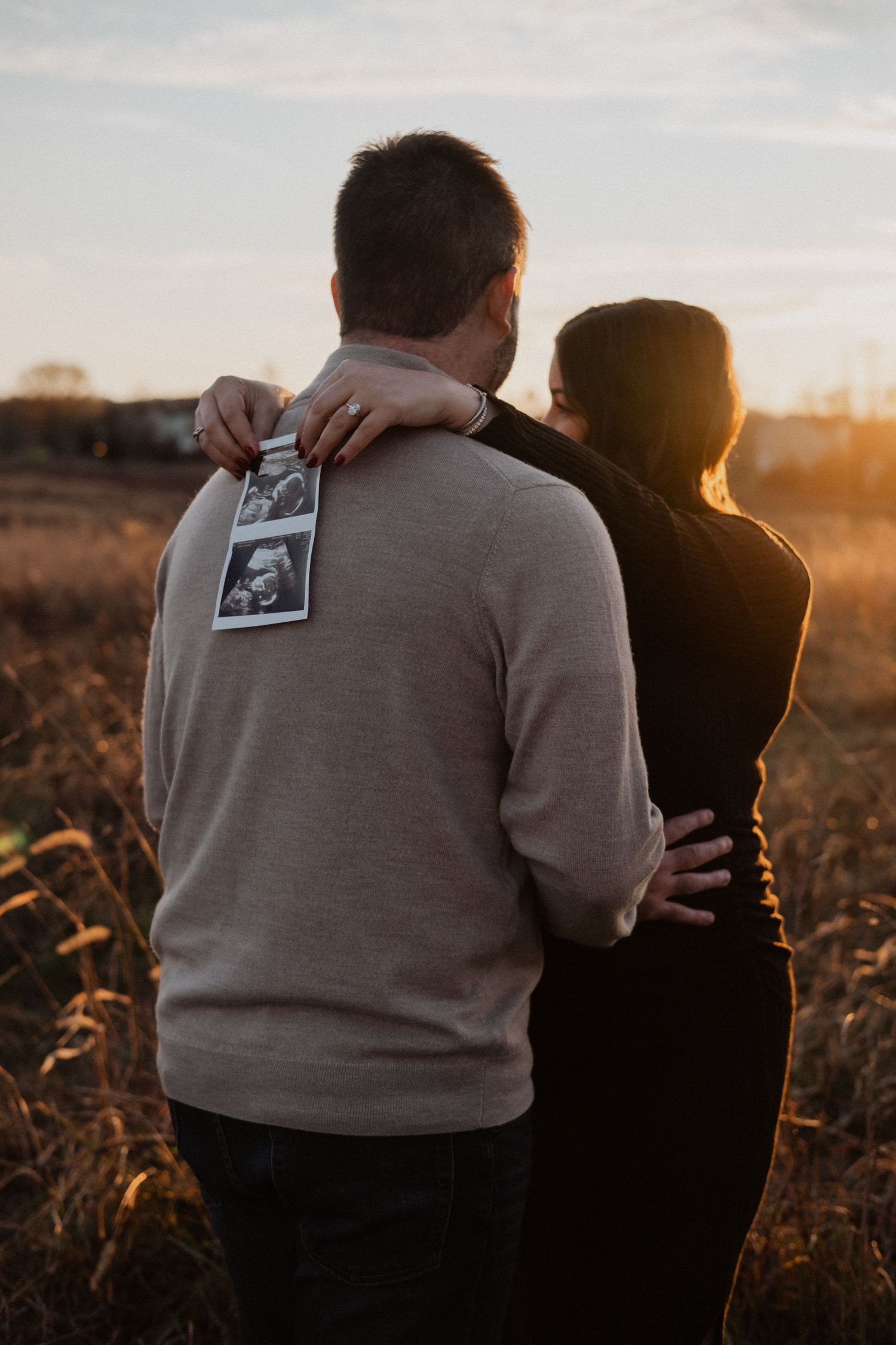 Couple embracing at sunset in a field, holding ultrasound photos, announcing pregnancy in golden hour light.