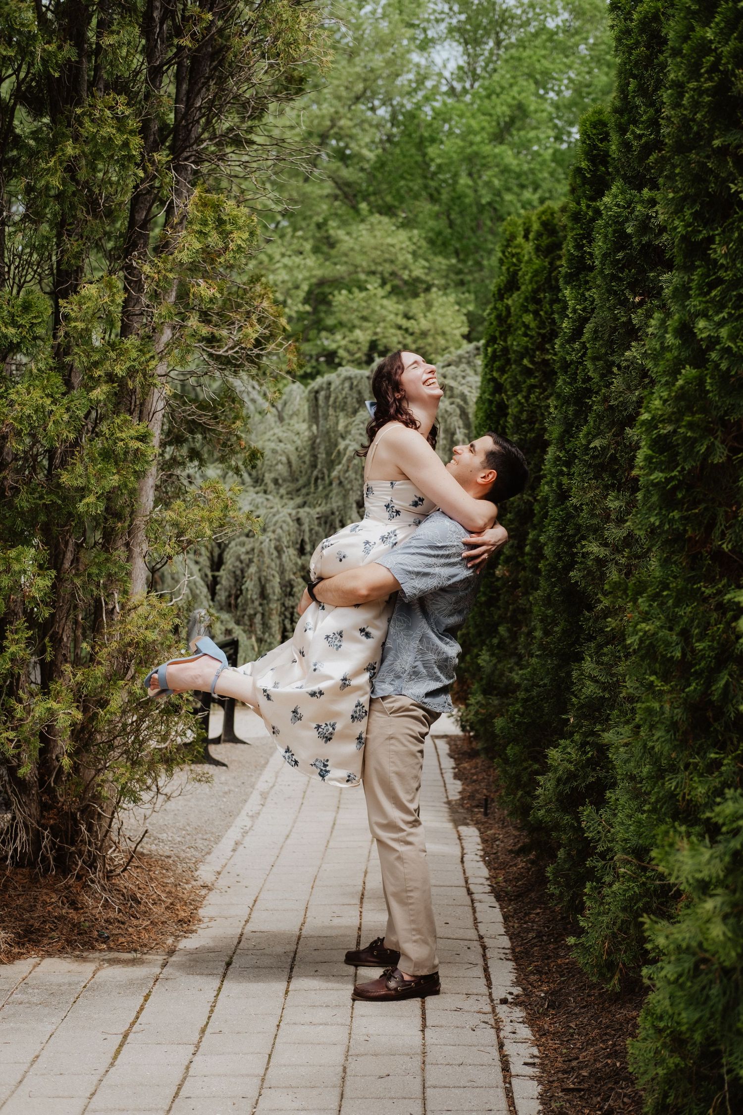 Joyful man lifting woman in floral dress along a tree-lined garden path, both laughing with happiness.