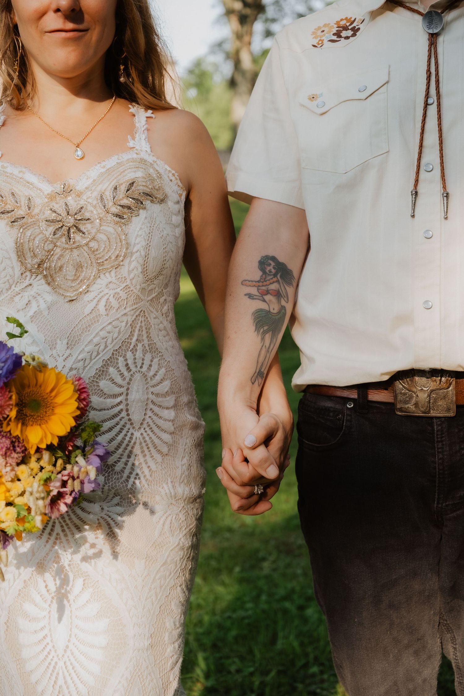 Bride in embroidered gown holding colorful bouquet holds hands with tattooed groom in western shirt.