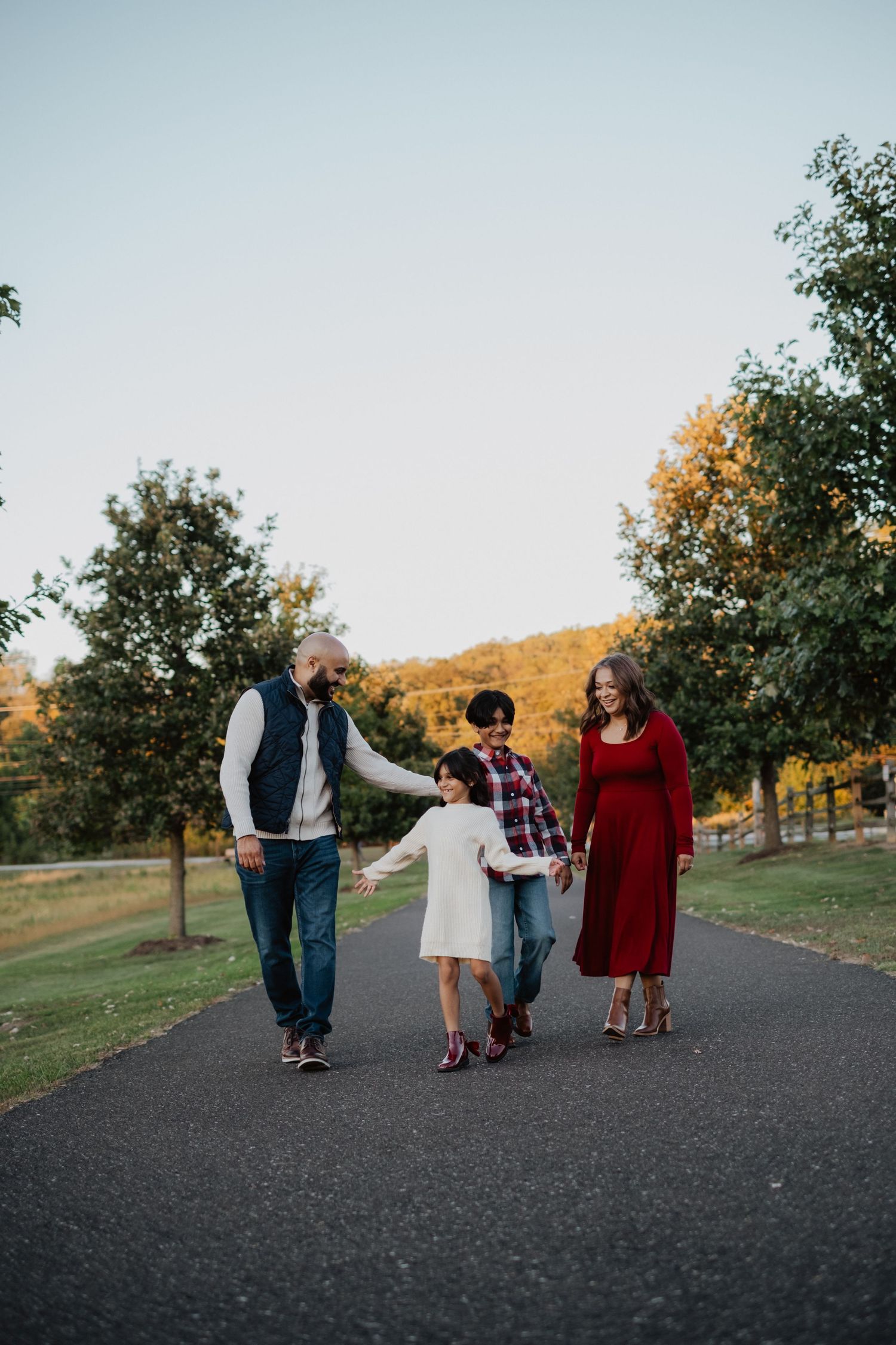 Family of four walking hand in hand on paved path surrounded by autumn foliage.