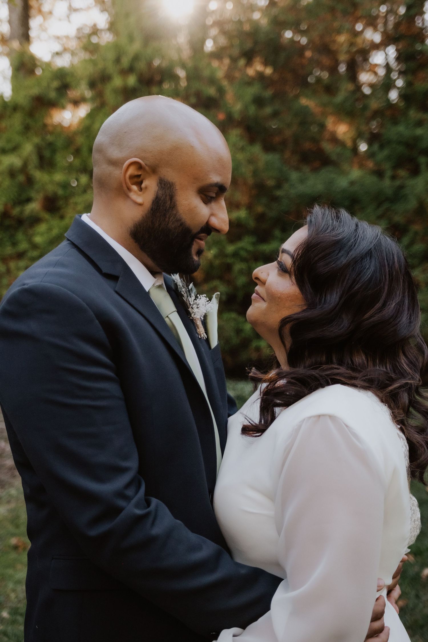 Bride and groom gazing at each other tenderly outdoors at sunset, dressed in white gown and navy suit.