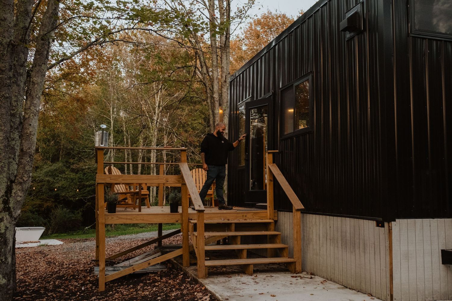Person standing on wooden deck of modern black cabin surrounded by autumn trees in a forest setting.