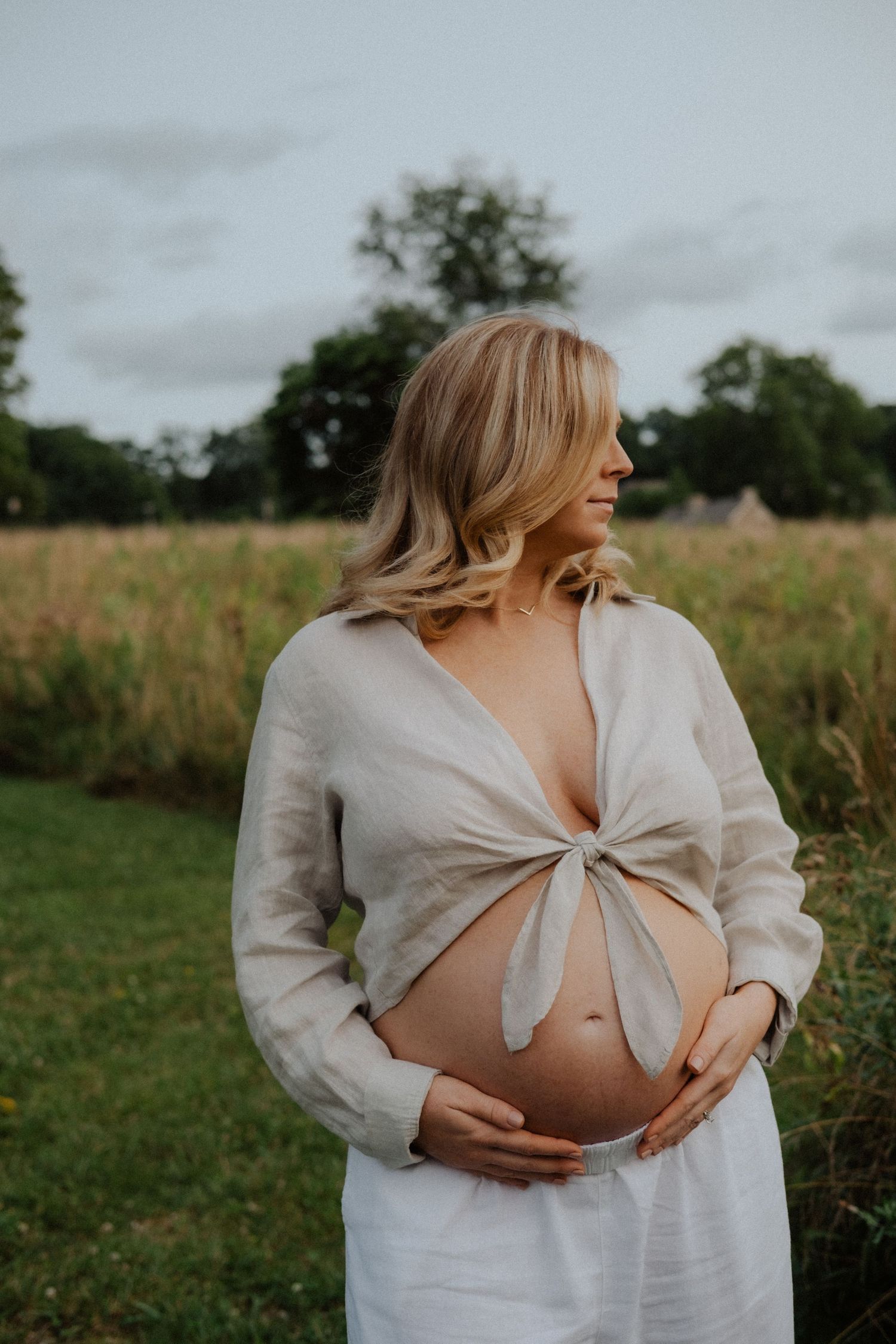 Pregnant woman in white linen top holding baby bump in a field.