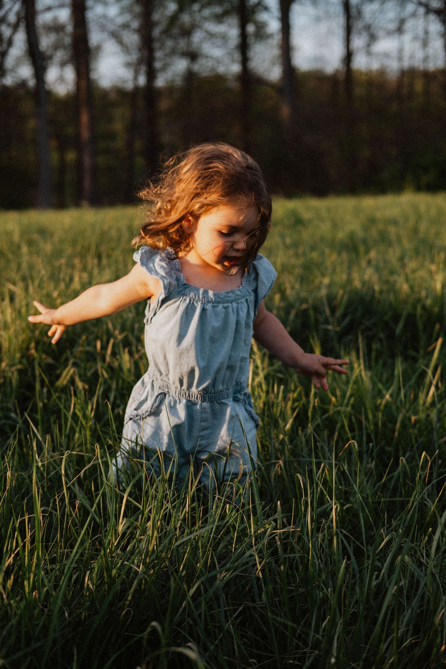 A young girl in a light blue dress runs through tall green grass at golden hour with trees in the background.