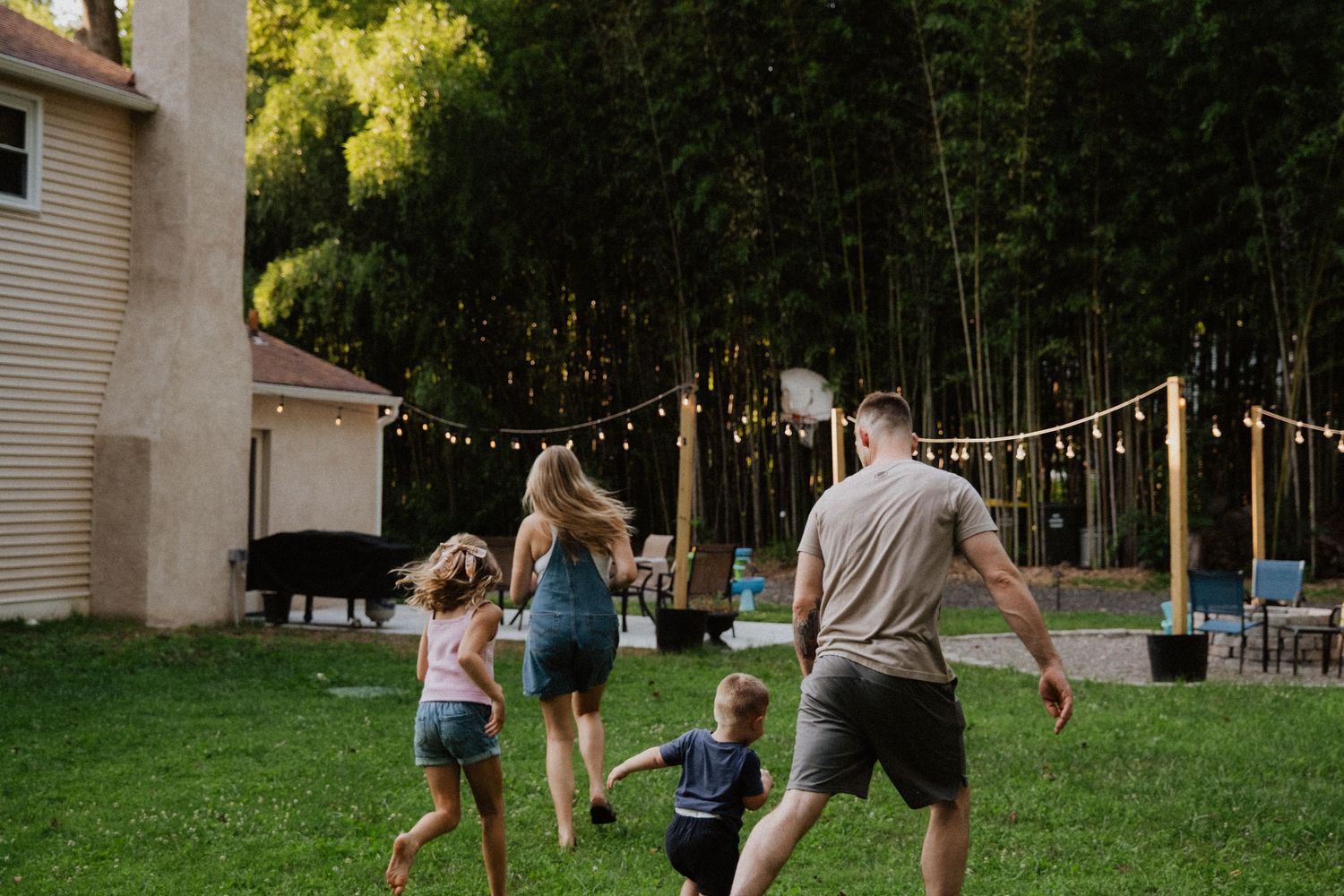 Family running and playing together in backyard with string lights and bamboo trees.