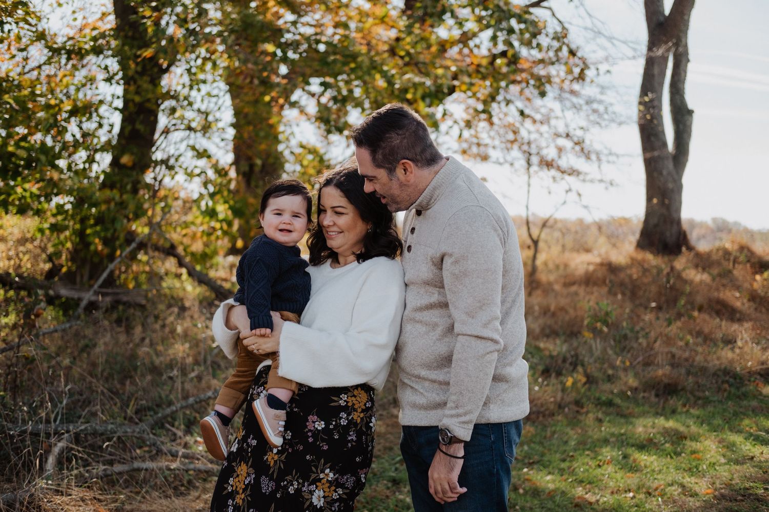 A happy family of three poses outdoors in autumn, parents smiling at their toddler among fall foliage.