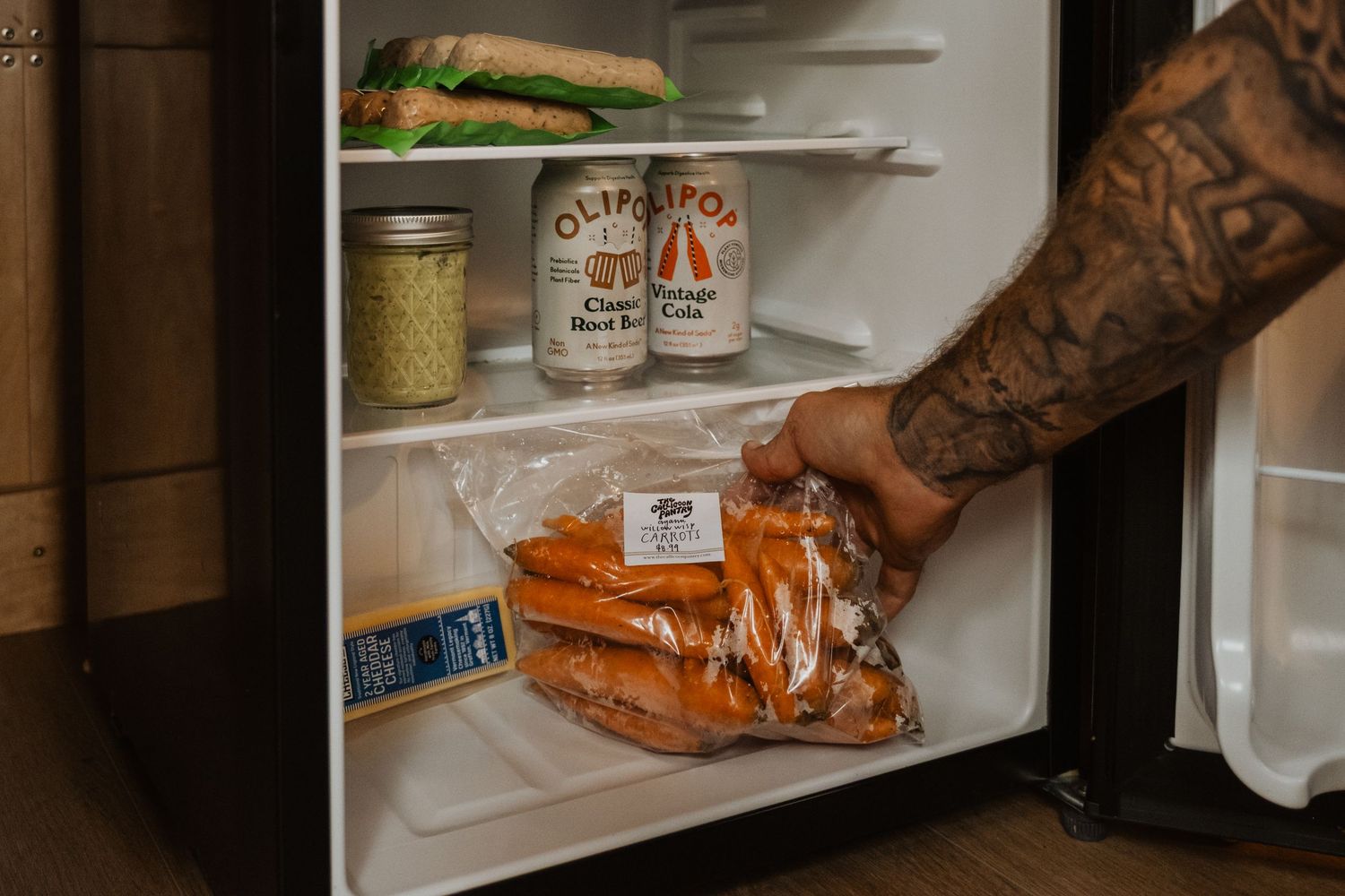 Tattooed hand placing a bag of carrots into a small refrigerator stocked with canned goods and a sandwich.