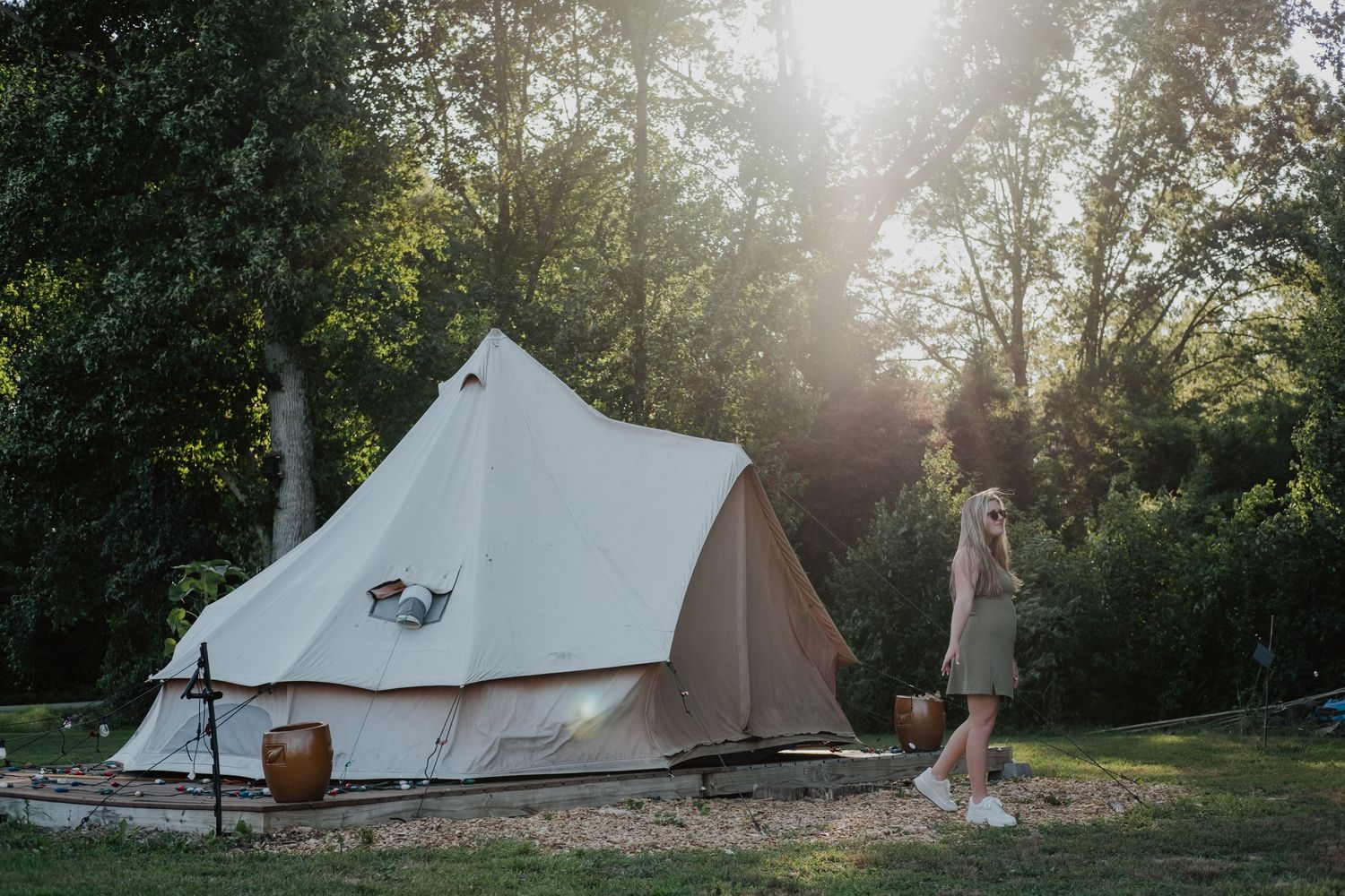 Woman in green dress standing beside a large canvas bell tent glamping setup in a lush green garden at sunset.