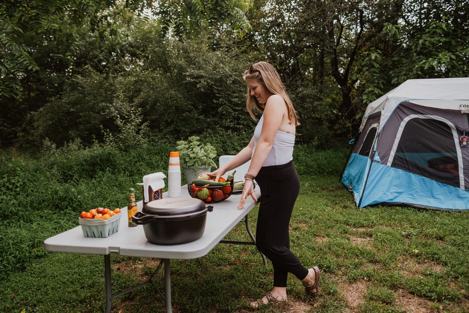 Woman preparing food at a camping table outdoors near a blue tent surrounded by lush green trees.