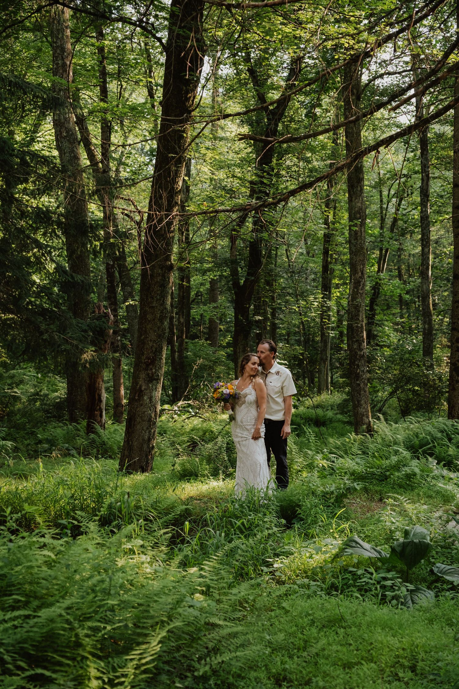 Bride and groom standing together in a lush green forest surrounded by ferns and tall trees.