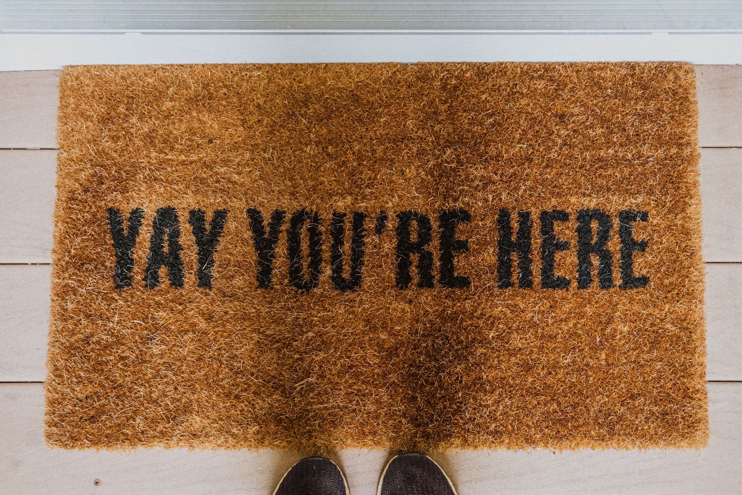 A welcome doormat reading YAY YOU RE HERE in bold black letters on natural coir with shoes visible at the bottom.
