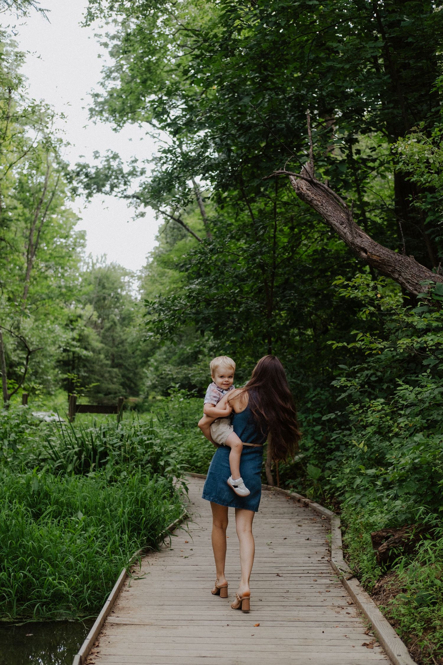Woman in denim skirt carries toddler on a wooden boardwalk path through lush green forest.
