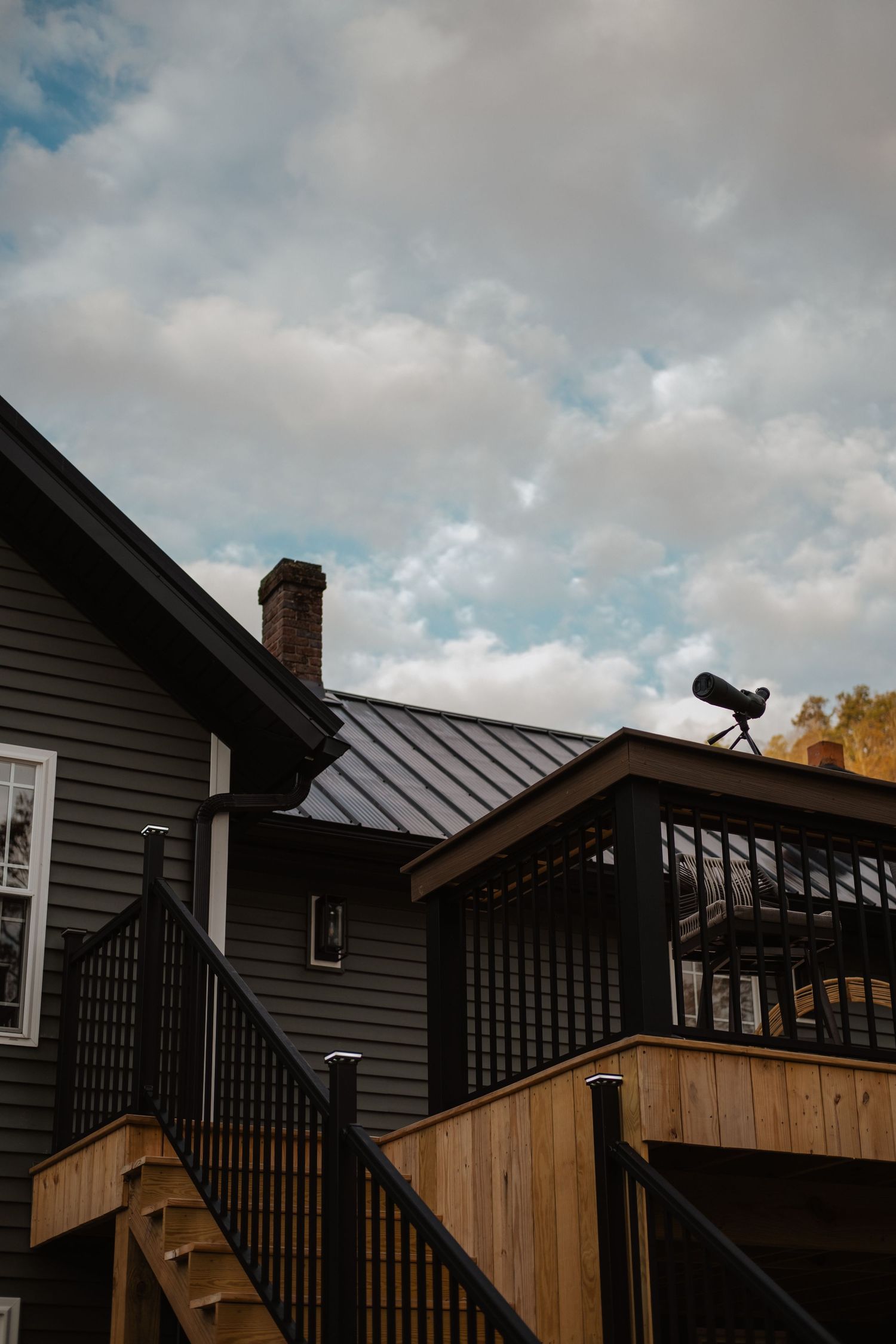 Dark gray house with metal roof, brick chimney, wooden deck stairs, and telescope under cloudy sky.