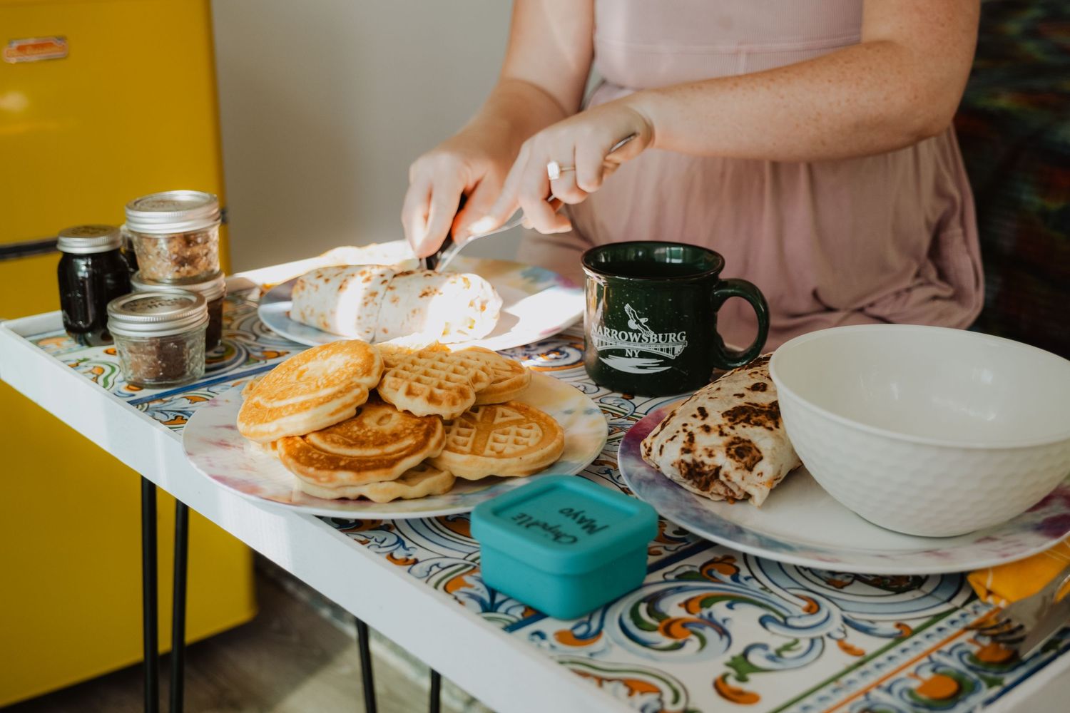 Woman spreading butter on bread at a table with waffles, pancakes, and a green mug for a hearty breakfast spread.