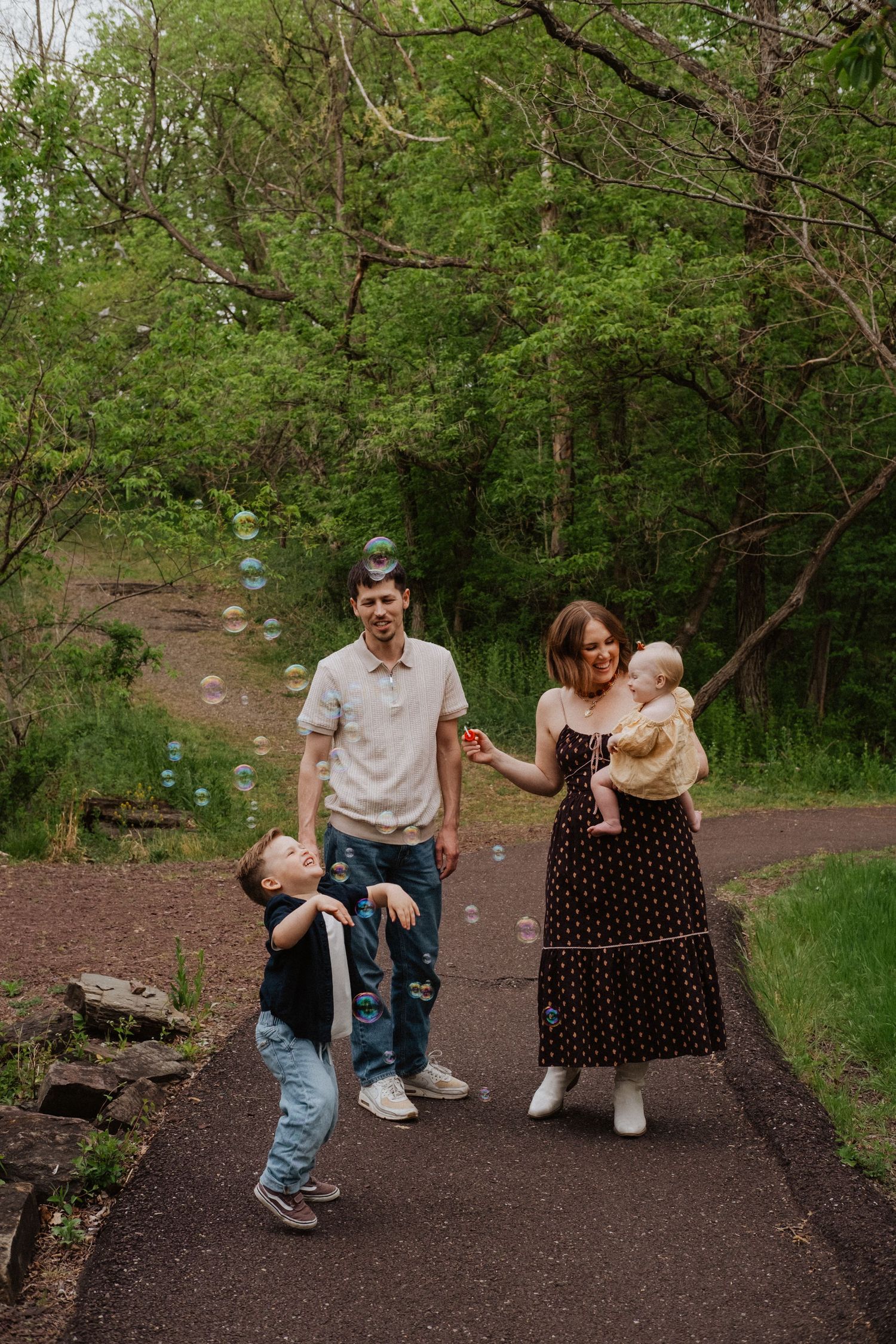 Family of four enjoying bubbles on a park path surrounded by lush green spring trees.